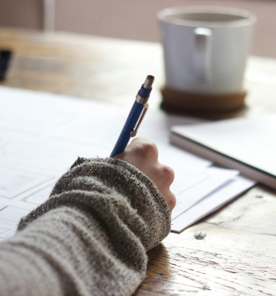 person writing on brown wooden table near white ceramic mug
